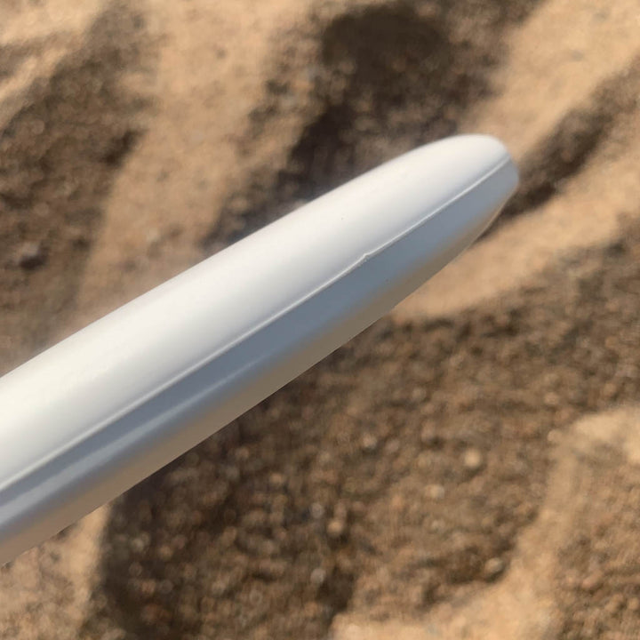 A close-up of the edge of a white Magic - Suregrip - SS disc golf putter designed for beginners, with a sandy surface in the background.