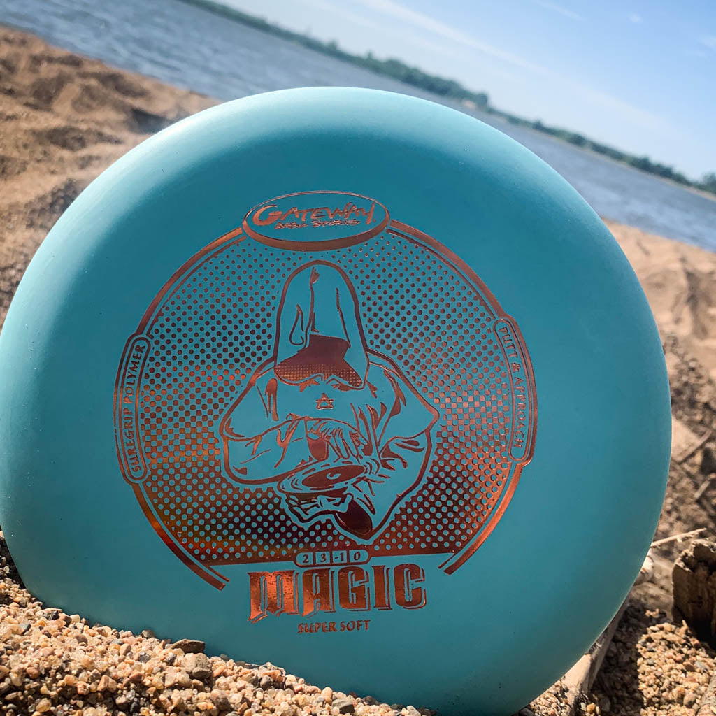 The Magic - Suregrip - SS is a blue, beginner-friendly putter with a hooded figure design on sandy ground by a lake, ideal for those who want a straight-flying disc.