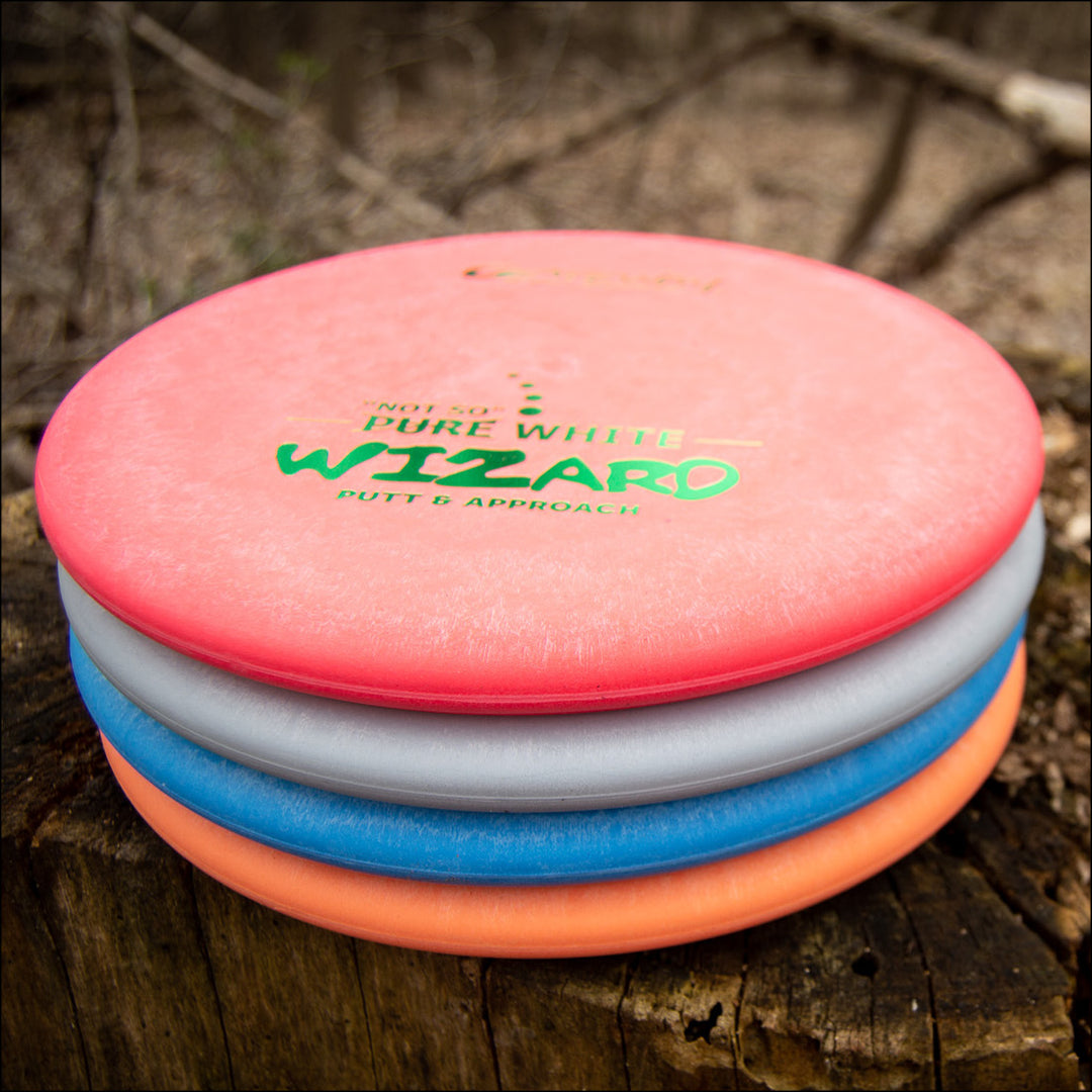 Four stacked Wizard - "Not So" Pure White disc golf putters rest on a tree stump in a wooded setting—perfect stable discs for accurate putts and reliable mid-range drives.