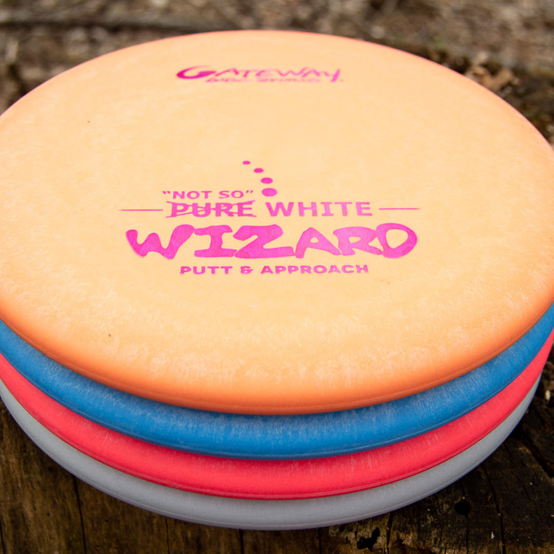 Stack of Gateway Wizard - "Not So" Pure White stable putters in orange, blue, and white on a tree stump—ideal for reliable mid-range drives and accurate putting.