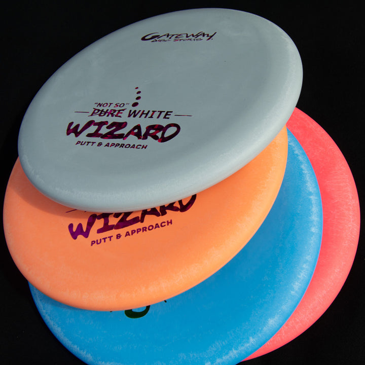 Four "Wizard - 'Not So' Pure White" disc golf putters, in white, orange, pink, and blue, are stacked on a black surface. This stable workhorse is ideal for consistent putts and short mid-range drives.