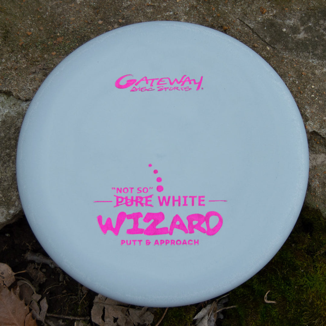 A Wizard - "Not So" Pure White disc golf putter with pink text, stable for putting and mid-range drives, lies on the ground—a reliable workhorse for disc golfers.