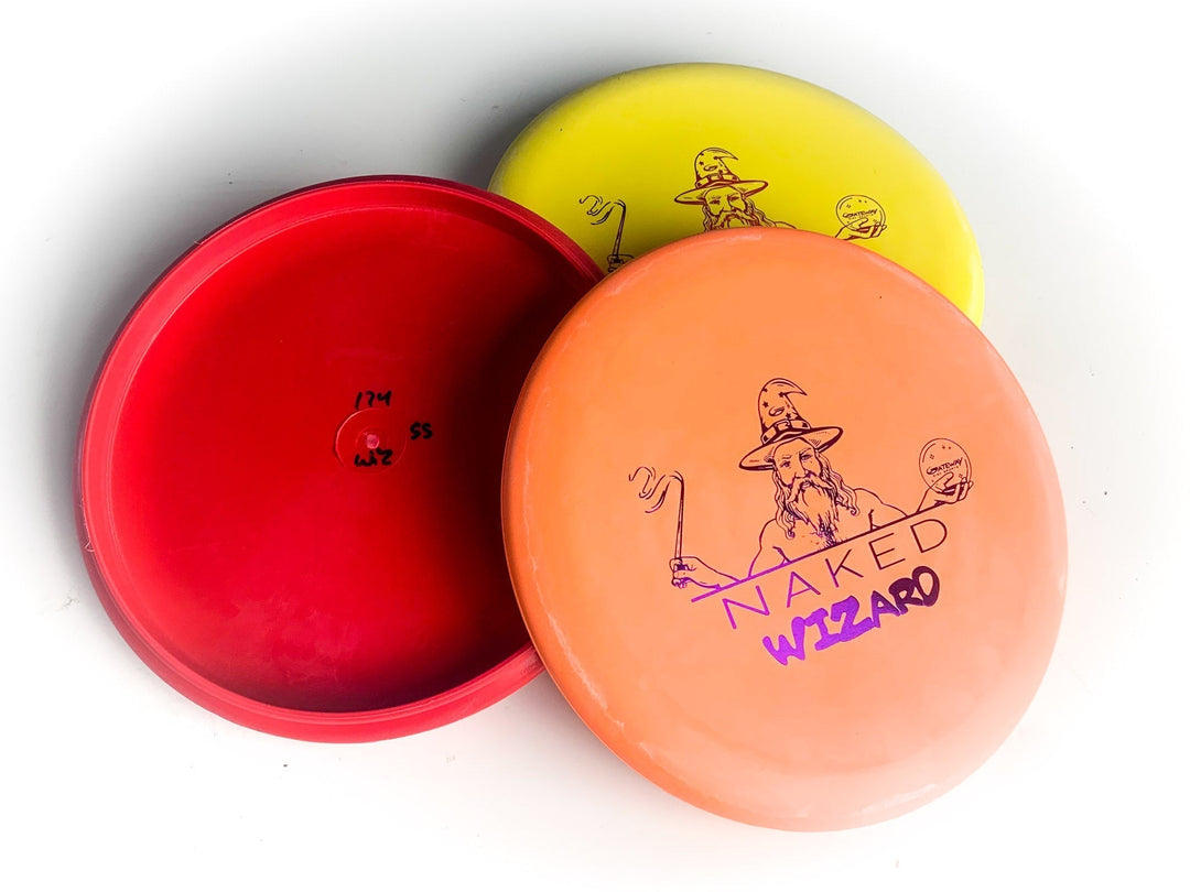 Three disc golf putters are shown: red and yellow discs with a wizard graphic and beaded putt design, plus a peach-colored Wizard - Firm - NAKED disc.