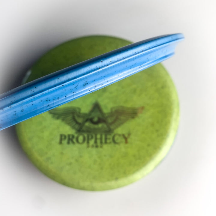 A blue disc partially rests on a green Prophecy - Diamond Hemp disc featuring an eye and wing logo, the word "PROPHECY," and is marked as an overstable mid-range for consistent flight.