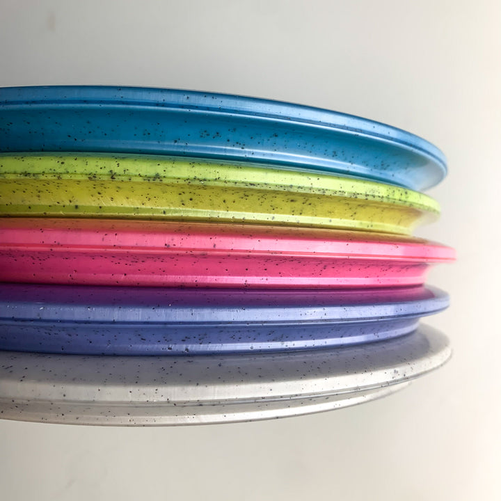 A close-up side view of six stacked Prophecy - Diamond Hemp plastic discs in blue, yellow, pink, purple, and white with speckled patterns, crafted for consistent flight, shown against a plain background.