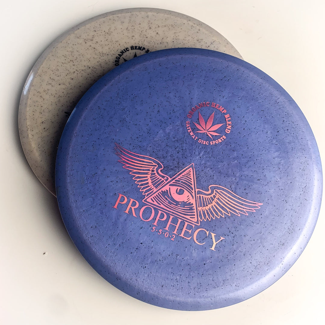 Two disc golf discs: one gray, one purple. The purple disc is the Prophecy - Diamond Hemp, featuring an eye with wings and a hemp leaf logo. Made of premium Diamond blend for overstable, consistent mid-range flight.