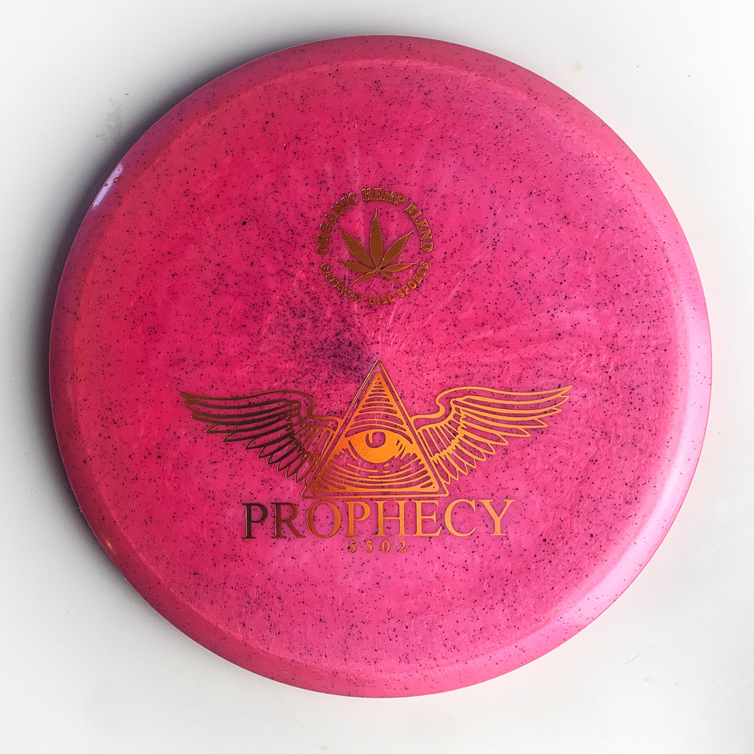 The Prophecy - Diamond Hemp is a pink disc golf disc with black speckles, made from premium Diamond blend for consistency. It features a gold winged eye triangle, "PROPHECY," and a leaf logo above.