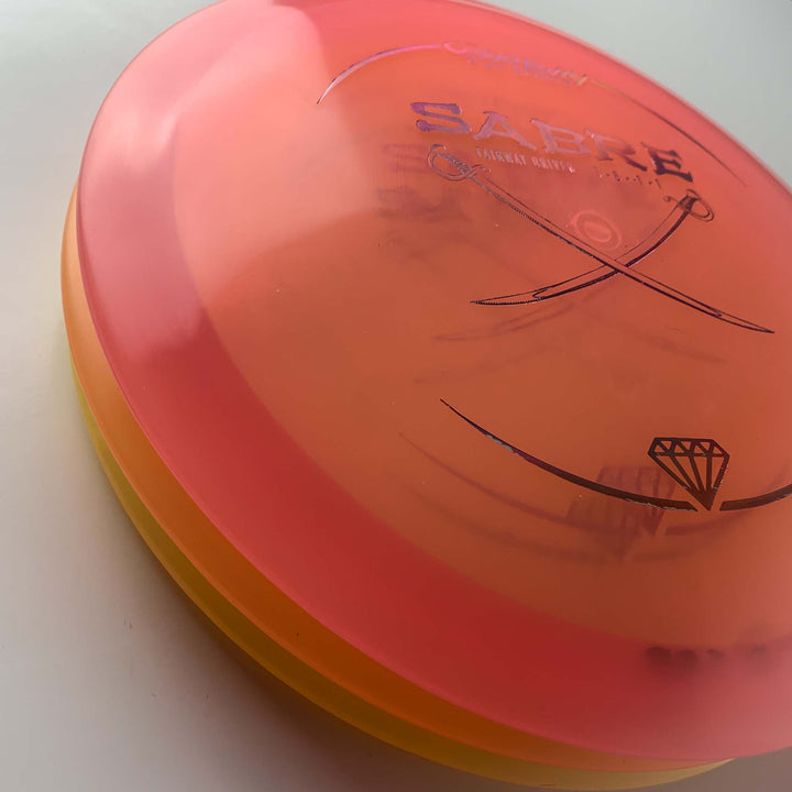 A close-up of the Sabre - Diamond disc golf disc, featuring red and orange colors with "SABRE" and saber sword graphics, known as one of the straightest drivers for its reliable, predictable finish.