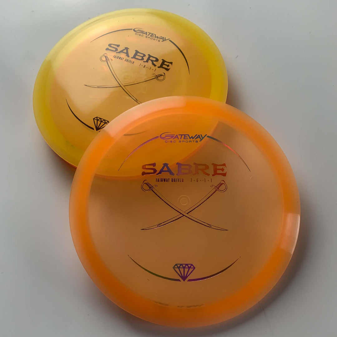 Two Sabre - Diamond disc golf discs, one yellow and one orange, are stacked on a white surface with the Sabre logo and crossed swords visible. The Sabre - Diamond is known for its versatile, straight driver flight and predictable finish.