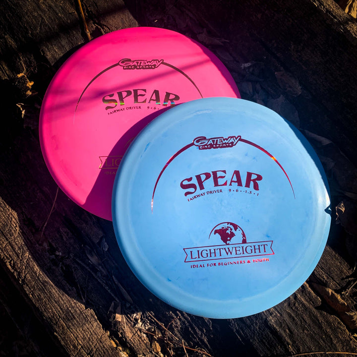A pink and a blue Spear - SureGrip Lightweight control driver disc golf disc rest on a wooden surface, sunlight highlighting their logos—perfect for accurate placement with every throw.
