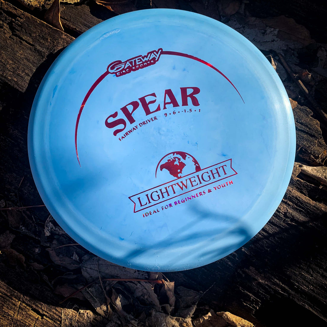 A blue Spear - SureGrip Lightweight control driver disc rests on dark soil scattered with fallen leaves.
