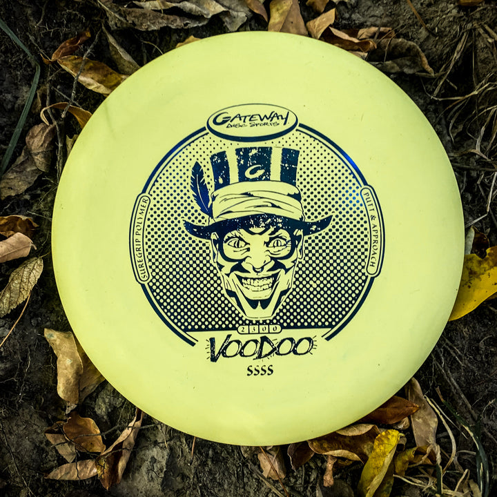 A yellow Voodoo - Suregrip - SSSS putter from Gateway Disc Sports lies on the ground among brown leaves, featuring a graphic of a grinning face in a top hat.