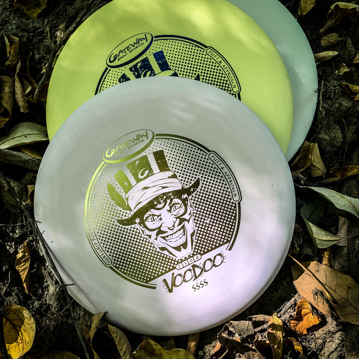 Two disc golf putters€”a white and a yellow "Voodoo - Suregrip - SSSS"€”each featuring a grinning character design, rest among dry leaves. Perfect as reliable putters or lay-up discs for precise shots on the course.
