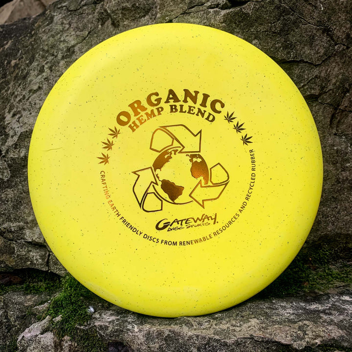 A yellow Warlock - Organic Hemp (H-SS) putter disc with an earth graphic and recycling symbol rests against a rock backdrop—made from organic hemp to enhance your approach game.