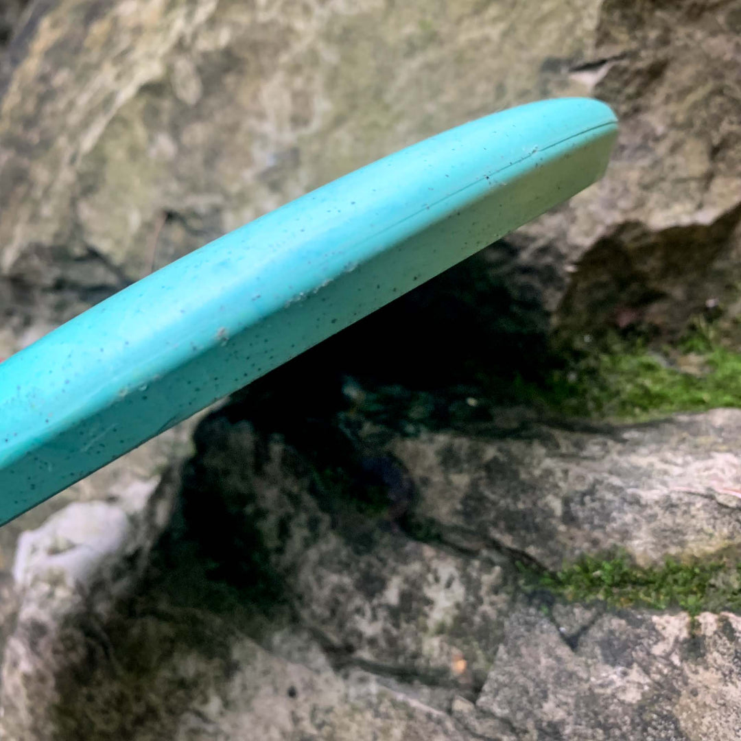 A close-up of the Warlock - Organic Hemp (H-SS) putter disc in light blue, made from organic hemp, resting on rocky terrain with green moss patches.