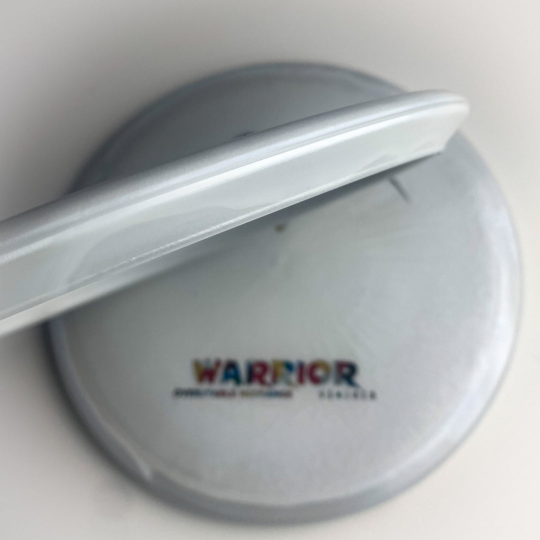 A close-up of the Warrior - Diamond Superglow, a white overstable mid-range disc with "WARRIOR" in colorful letters and smaller text below on its circular surface.