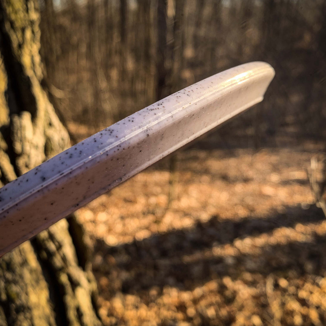 A close-up of the edge of the Warrior - Organic Hemp speckled mid-range disc golf disc, set against a blurred sunlit forest background.