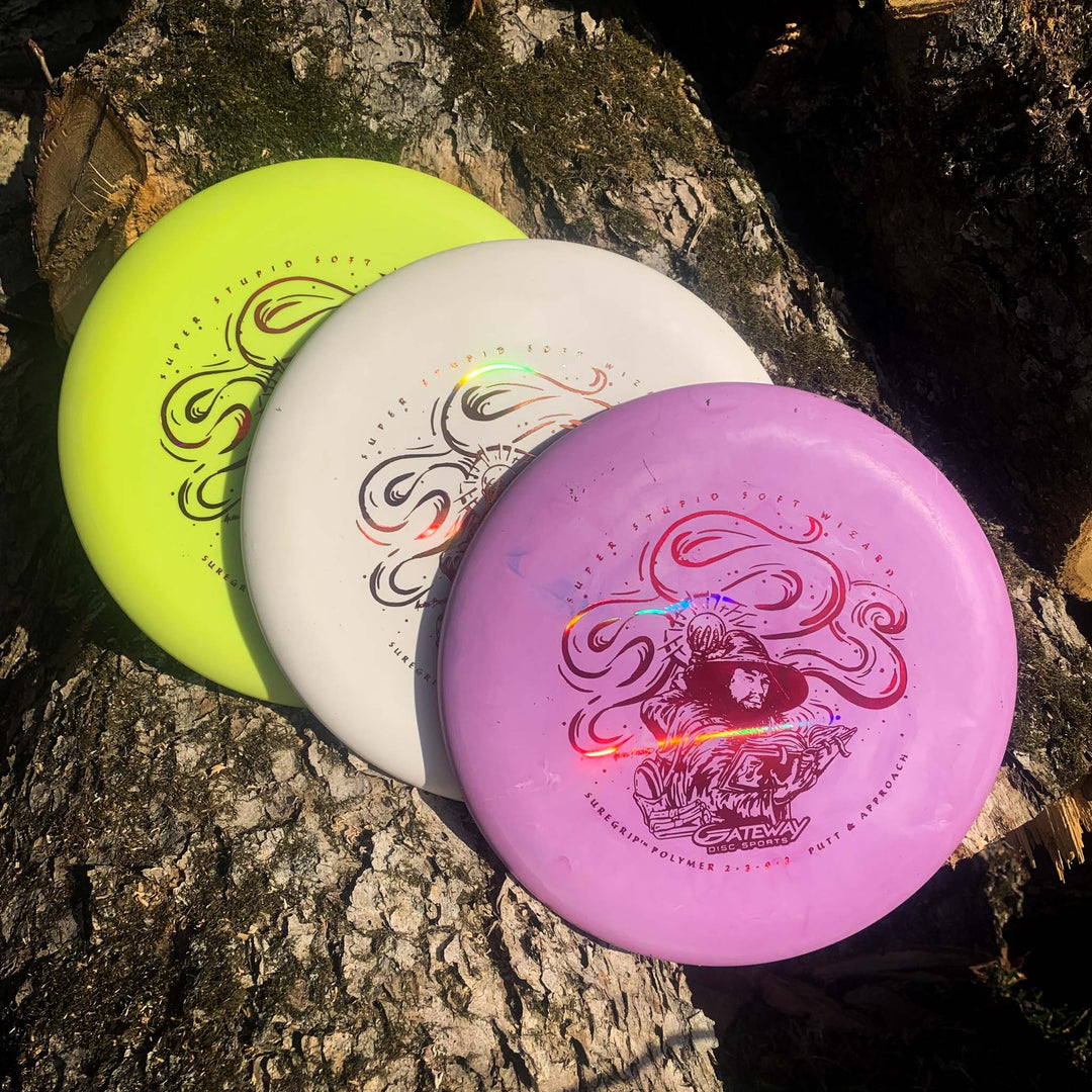Three Gateway Wizard - SSS disc golf discs in yellow, white, and purple rest on a tree trunk, each featuring a detailed mushroom-themed graphic. Ideal for accurate tricky shots or as a stable putter in windy conditions.