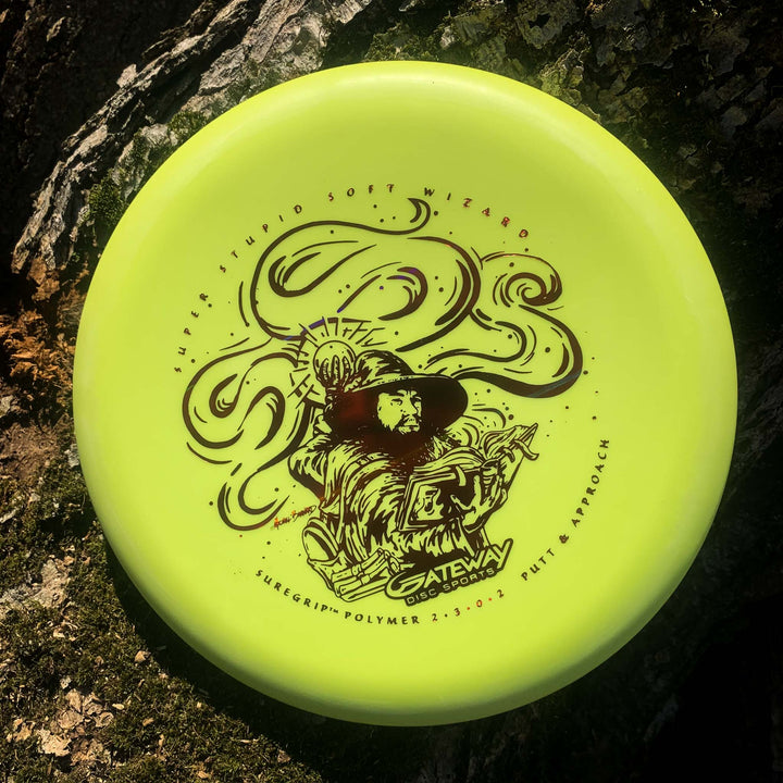 A yellow Wizard - SSS putter, featuring a black wizard with staff illustration and "Gateway" branding, rests on tree bark—ideal for stable putts and accurate throws, even in the wind.