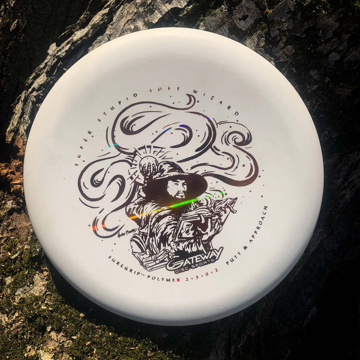 The Wizard - SSS, a white stable putter by Gateway with a wizard graphic, sits on rough tree bark in sunlight—perfect for precise throws even in windy conditions.