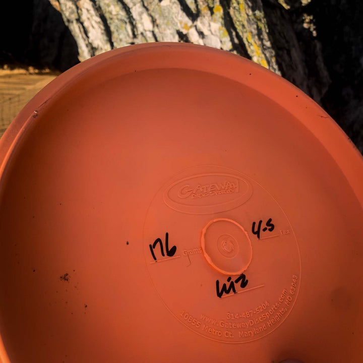 Close-up of the underside of an orange Wizard - SSSS putt and approach disc, featuring handwritten numbers and letters, with tree bark in the background.