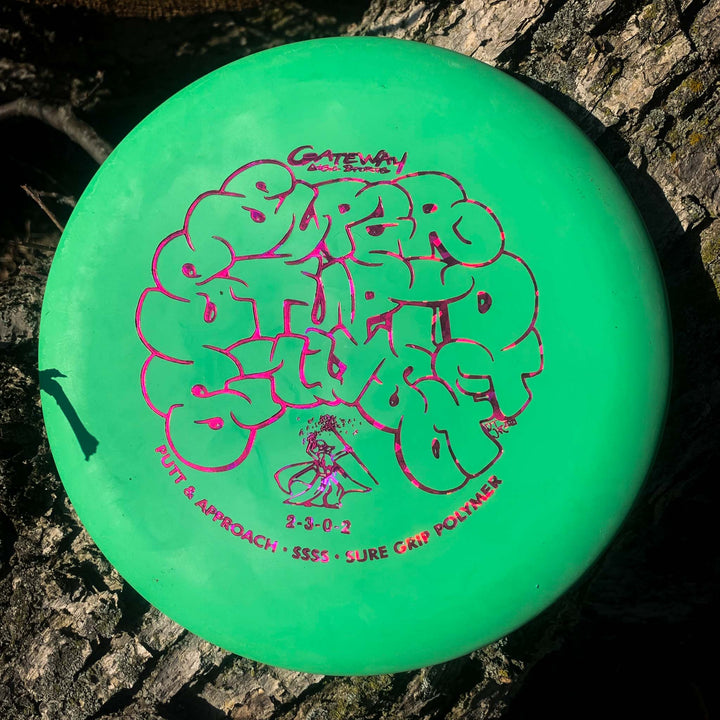 A Wizard - SSSS putt and approach disc with Gateway branding, featuring a green color and "Super Stupid Soft" plastic, sits on tree bark in sunlight.
