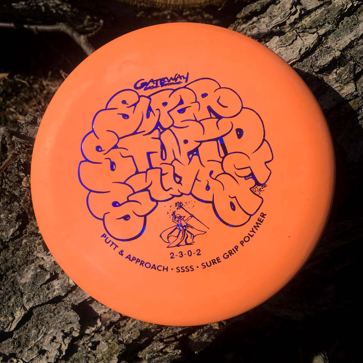 An orange Wizard - SSSS, a beaded putt and approach disc, lies on rough tree bark.