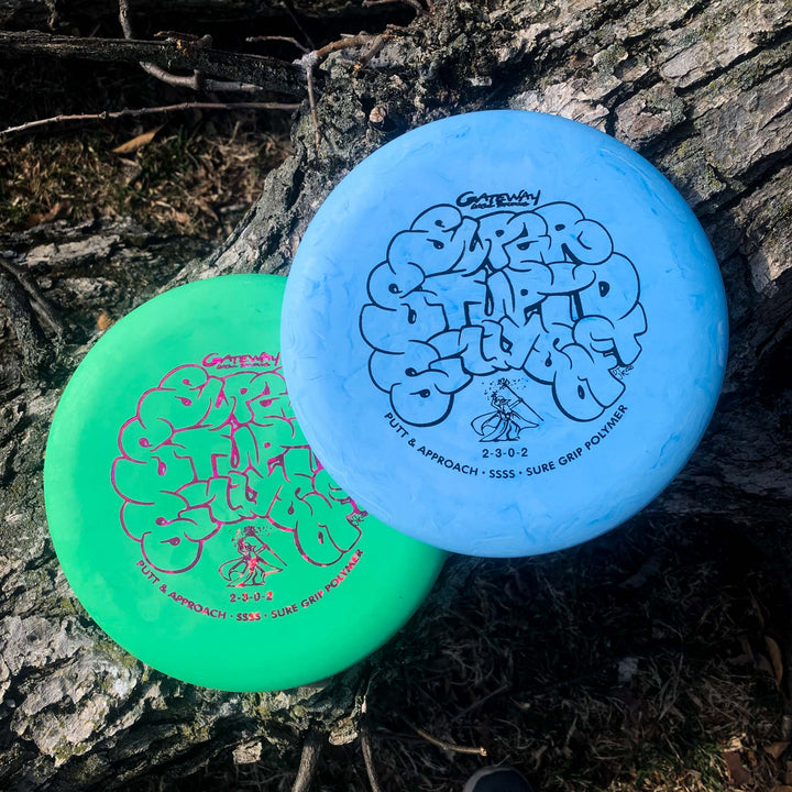 Two Wizard - SSSS disc golf putters, one green and one blue, rest on a textured tree trunk outdoors. These beaded putt and approach discs offer reliable performance for short throws.
