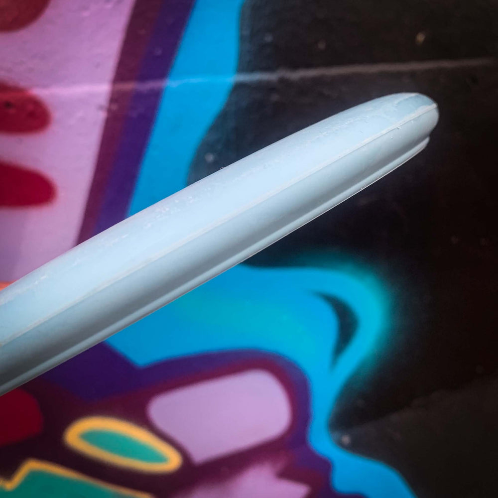 Close-up of the edge of a light blue Wizard - Suregrip Nylon putter frisbee in front of a colorful graffiti wall with blue, purple, and red shapes.