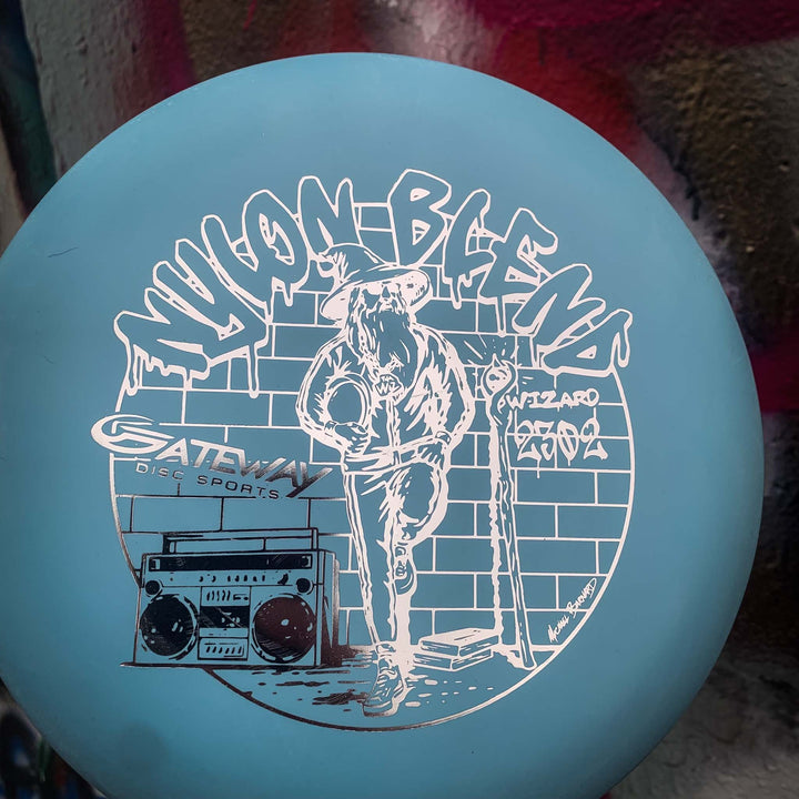 The Wizard - Suregrip Nylon is a blue disc golf disc from Gateway Disc Sports, featuring a wizard with a staff and boombox, plus graffiti "WIZARD" text. Ideal as a stable putter for confident mid-range drives.