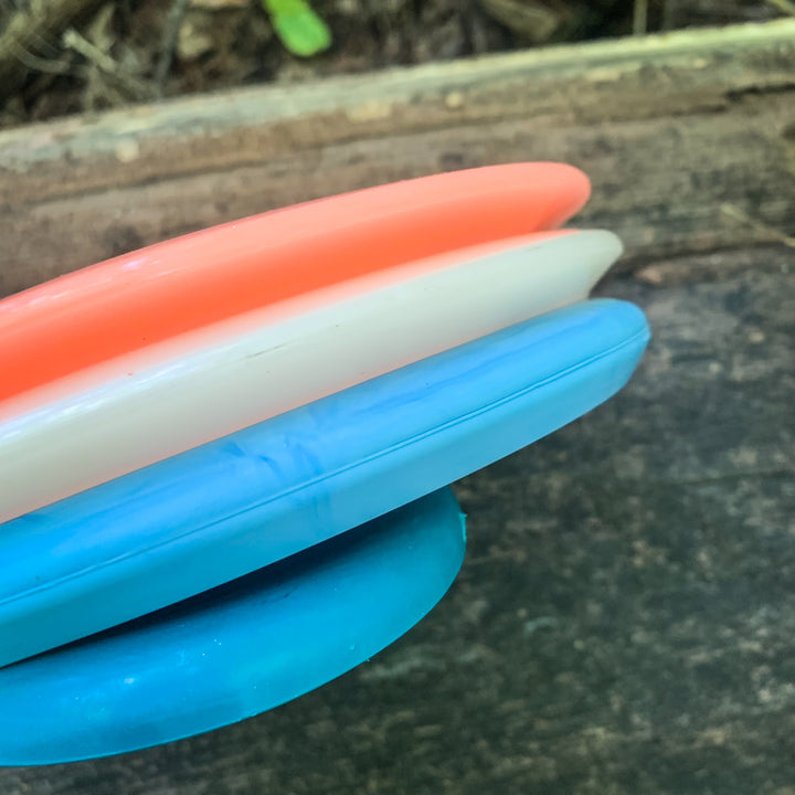 Close-up of a Factory 2nd 3-Disc Starter Pack with orange, white, and blue discs stacked on a wooden surface outdoors.