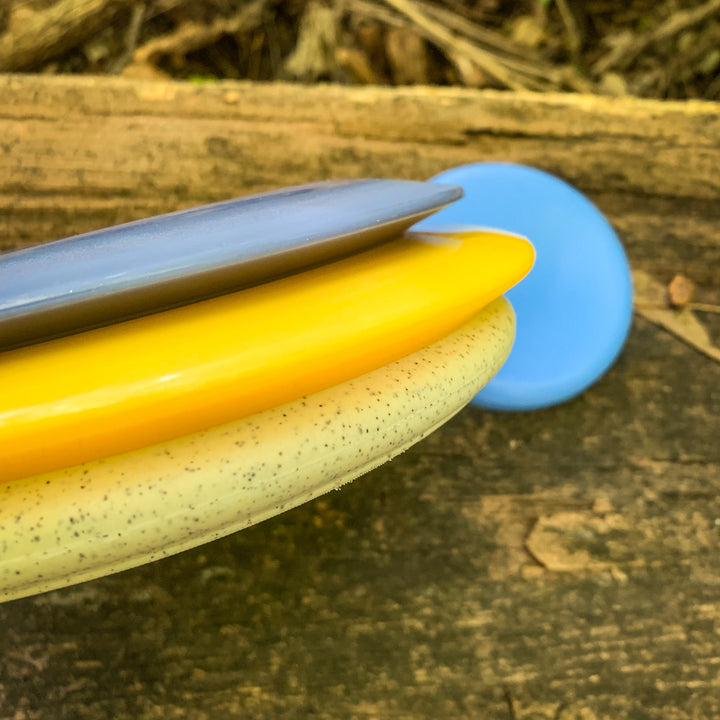 A Factory 2nd 3-Disc Starter Pack—featuring gray, yellow, and white speckled discs, including a SureGrip putter—stacked on wood, with a blue disc set apart in the background.