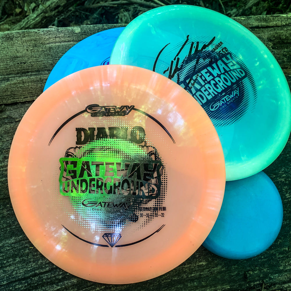 A Factory 2nd 3-Disc Starter Pack—featuring a peach "Diablo" Premium driver and two blue-green "Gateway Underground" SureGrip putters—rests on a wooden surface outdoors.