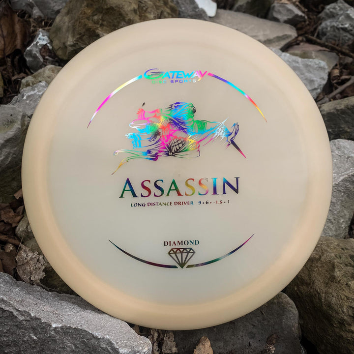 A cream-colored Assassin - Diamond distance driver by Gateway Sports rests on gray rocks, showcasing a colorful graphic and diamond logo, renowned for its excellent glide.