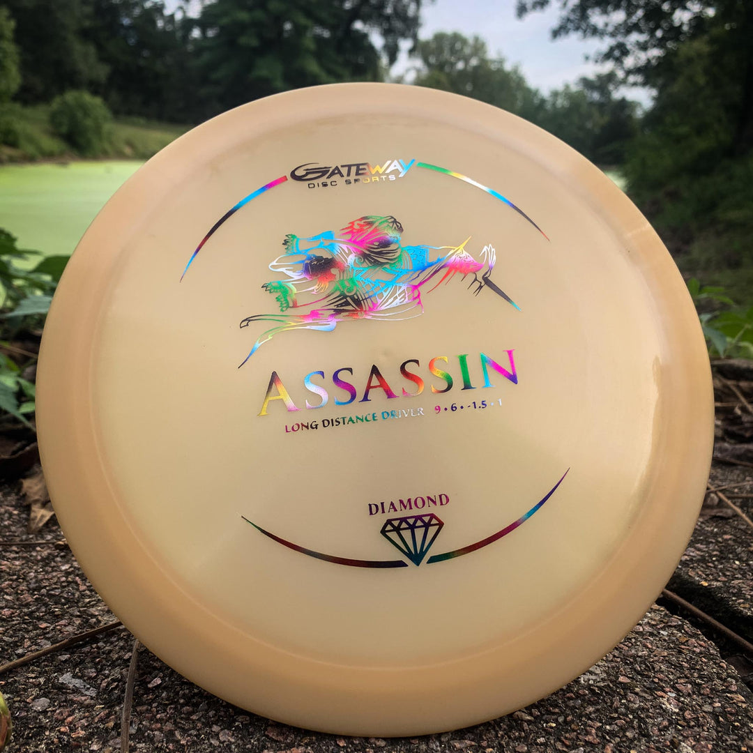 A Gateway Assassin - Diamond distance driver disc with a holographic logo and diamond symbol lies on the ground outdoors. Renowned for its neutral flight path and excellent glide.