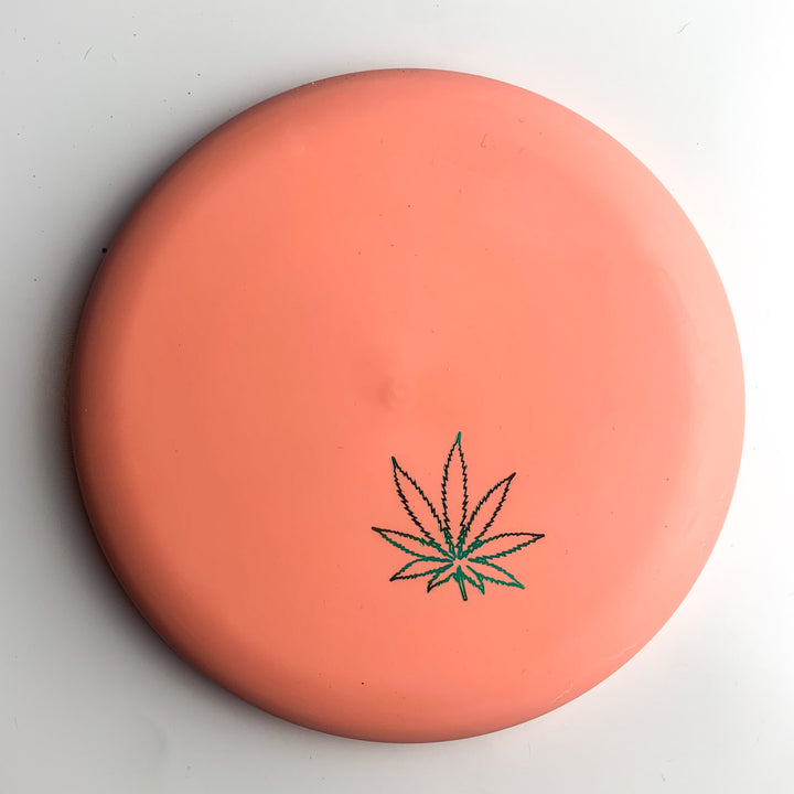 On a white surface rests the Wizard Leaf Suregrip Putter from Gateway Disc Sports—a round, orange disc with the iconic leaf graphic at its center.