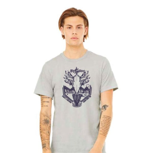 A young person with straight brown hair wears the Big $ T-Shirt€”light grey cotton with Ivan Marston's artwork€”standing against a plain white background, tattoos visible on both forearms.