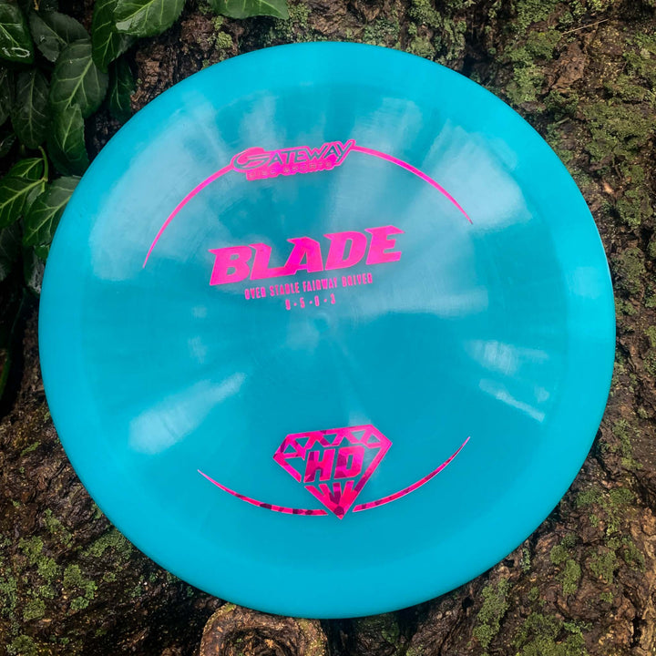A turquoise Blade - HD - NOT CURRENT VERSION overstable fairway driver with pink text and a diamond graphic leans against tree bark and foliage, offering reliable resistance and a consistent finish on the course.