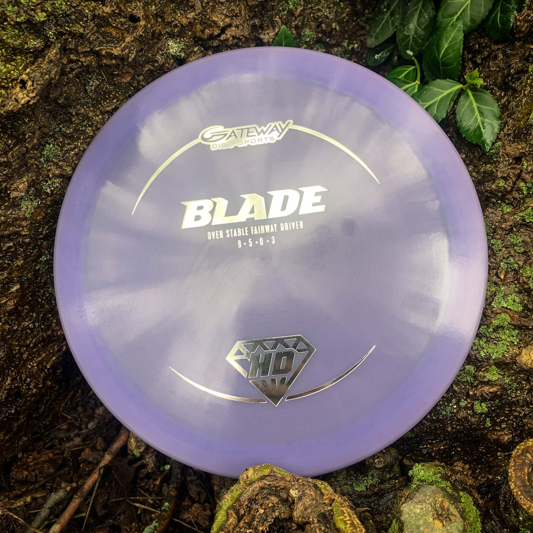 A Blade - HD - NOT CURRENT VERSION fairway driver by Gateway Disc Sports, in purple, rests on a mossy tree trunk beside green leaves, providing reliable overstability and a consistent finish.