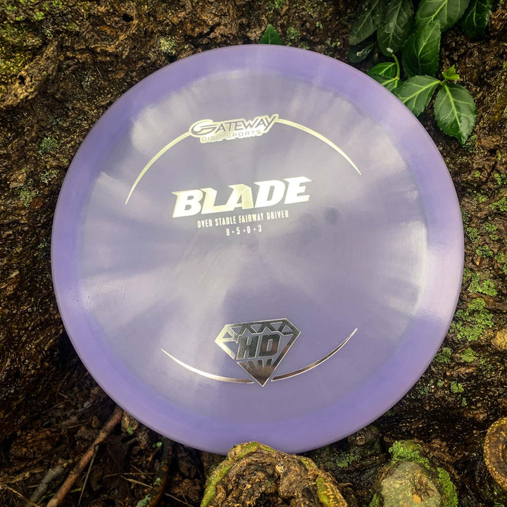 A Blade - HD - NOT CURRENT VERSION fairway driver by Gateway Disc Sports, in purple, rests on a mossy tree trunk beside green leaves, providing reliable overstability and a consistent finish.
