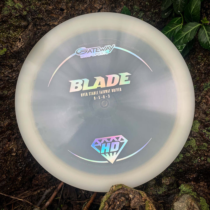 The Blade - HD - NOT CURRENT VERSION, an overstable fairway driver featuring "Blade" and "HD" logos, lies near leaves and roots, offering steady resistance and a reliable finish for your disc golf rounds.