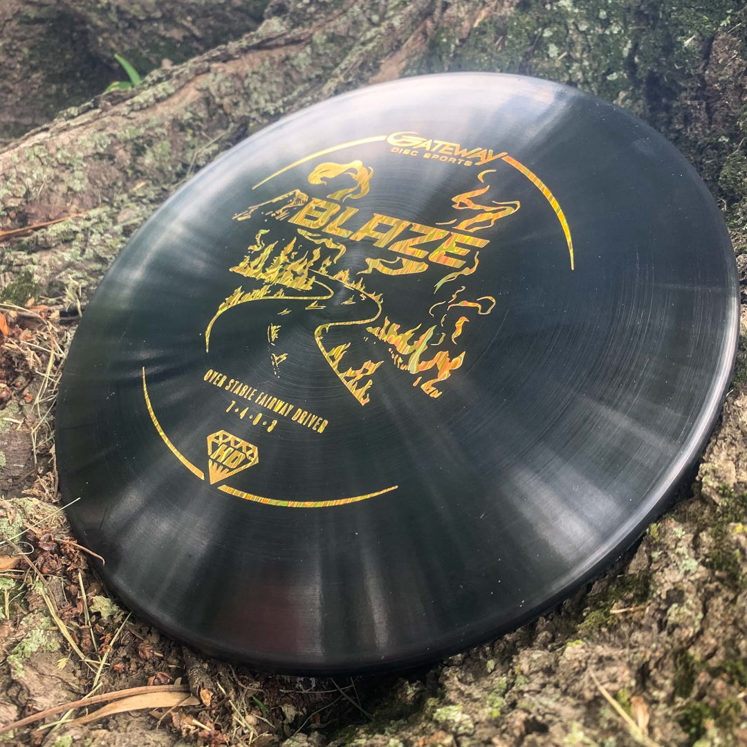 A black Blaze - Hyper Diamond (HD) overstable driver with gold graphics rests on tree roots outdoors, ready to deliver a consistent, predictable drive even in headwinds.