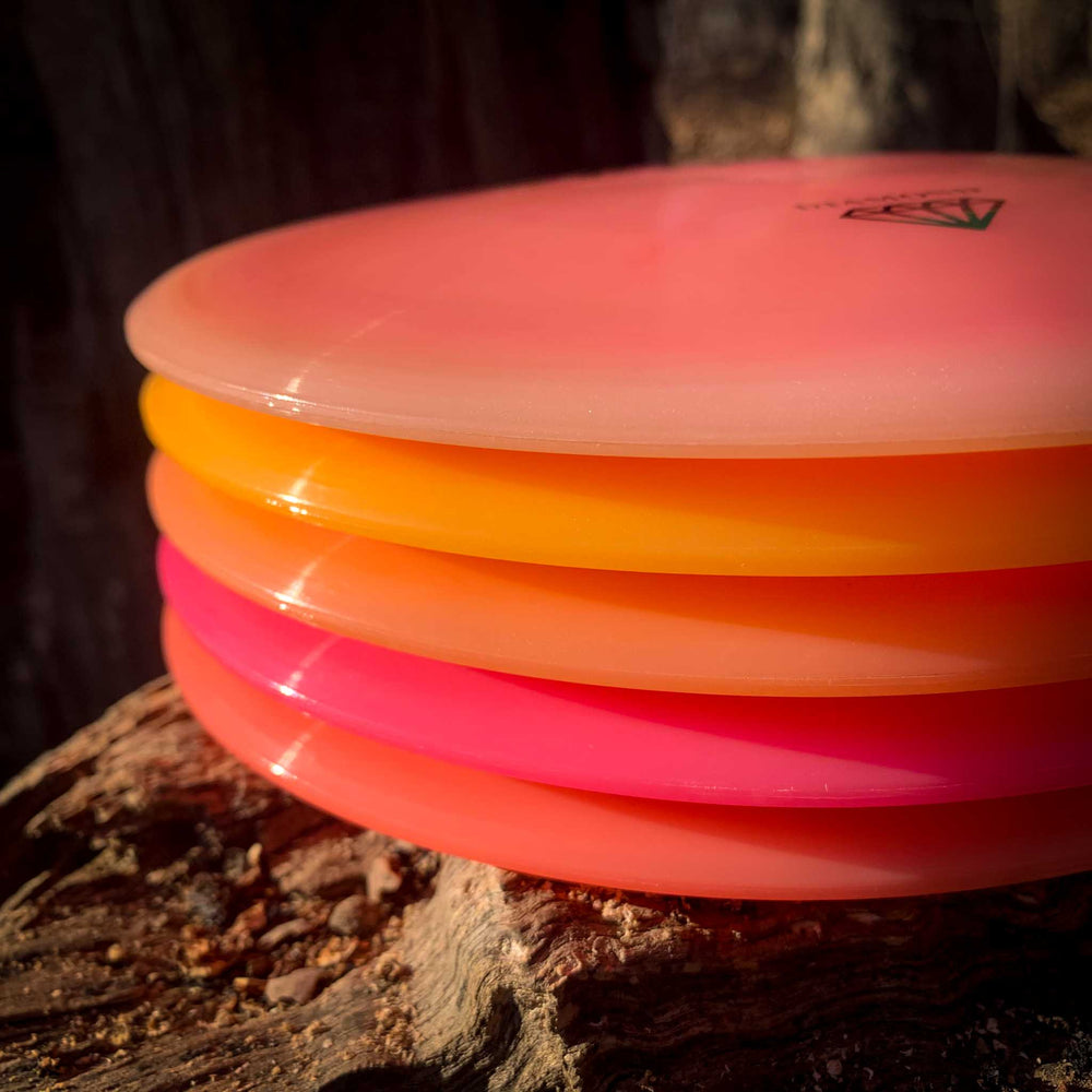 A stack of brightly colored Spirit - Diamond disc golf discs in pink, orange, and yellow rests on a piece of wood outdoors.
