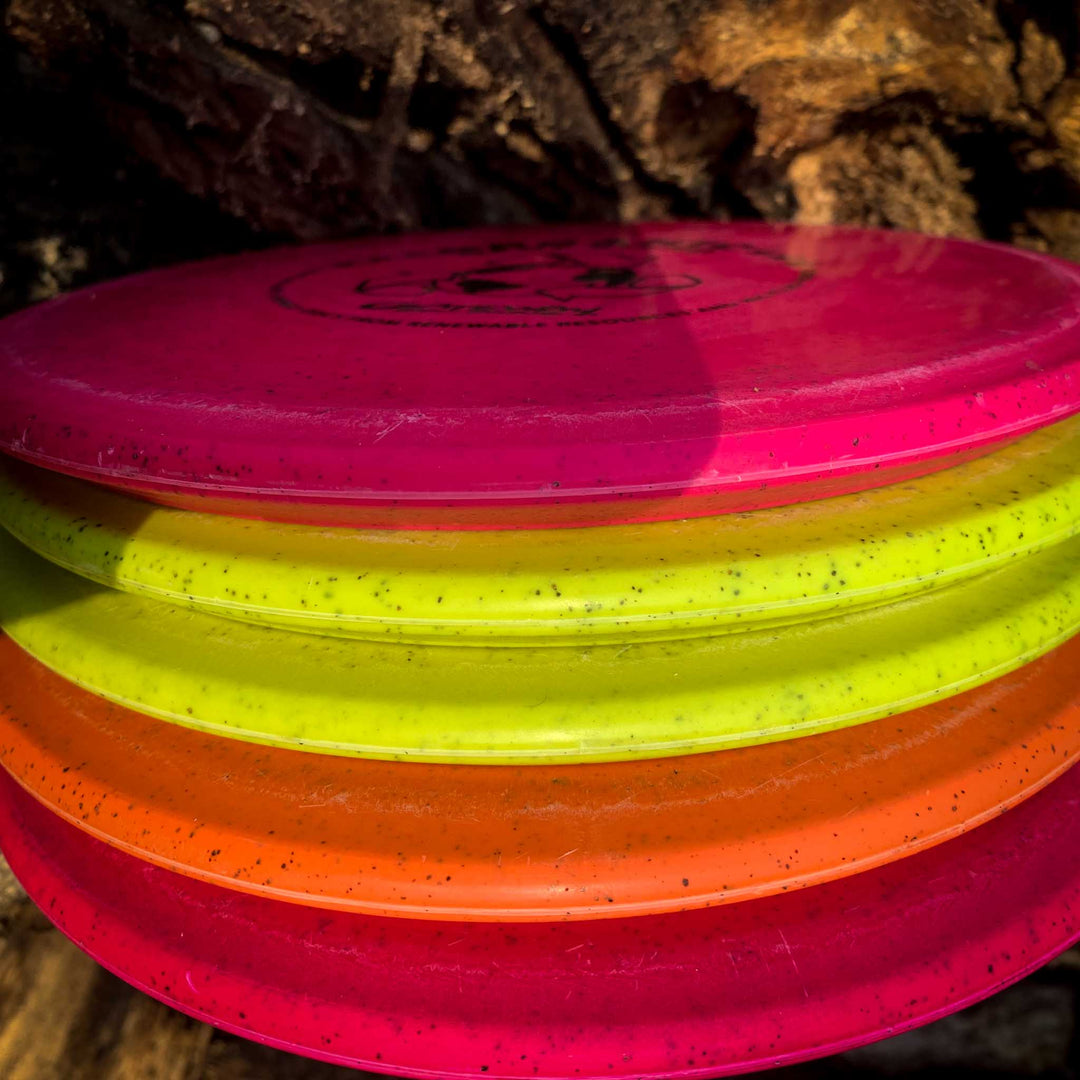 A close-up of four stacked Warrior - Organic Hemp disc golf discs—an overstable mid-range—in pink, yellow, orange, and red, with a blurred tree trunk in the background.