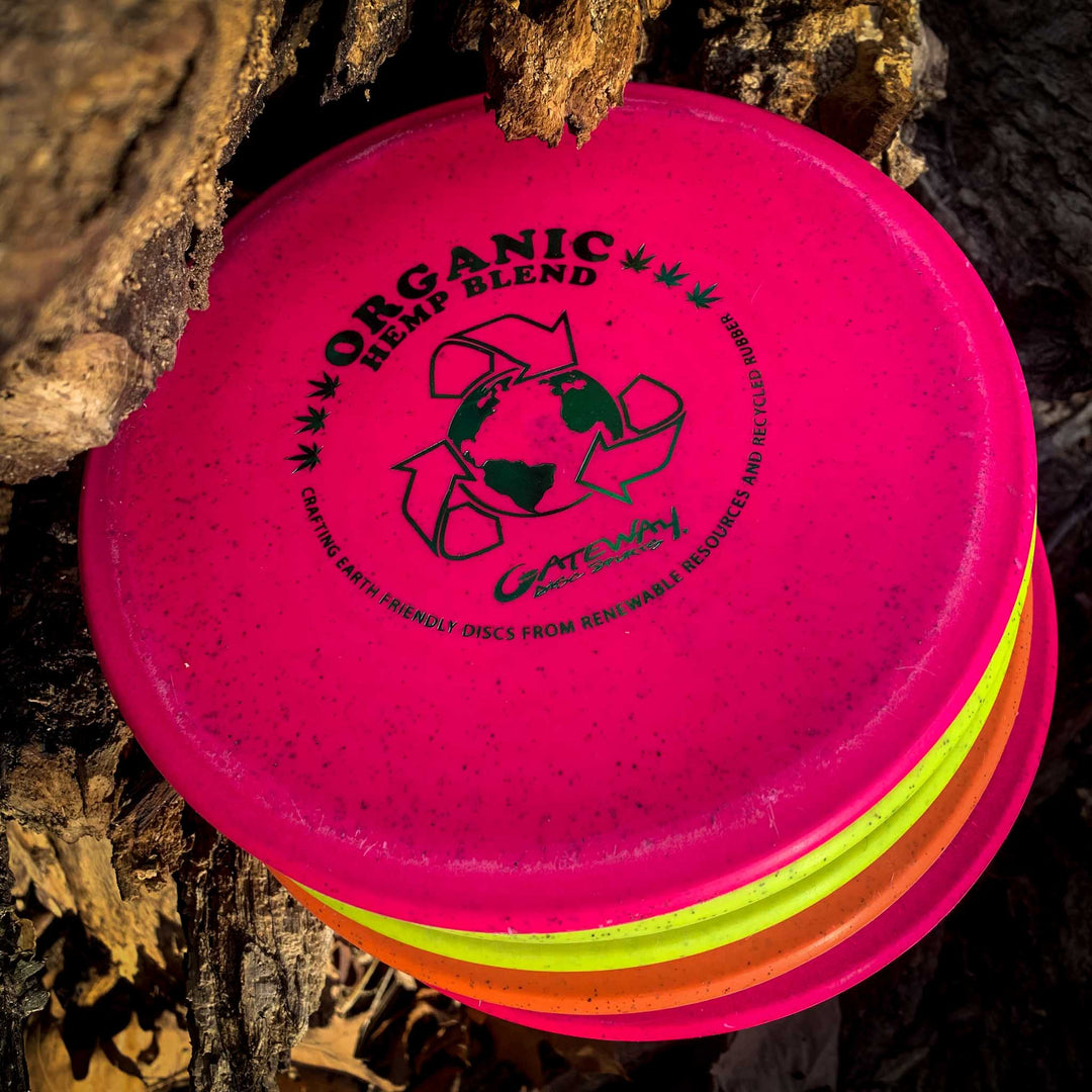 A stack of vibrant disc golf discs, featuring the Warrior - Organic Hemp mid-range with an environmental recycling logo, rests against tree bark.