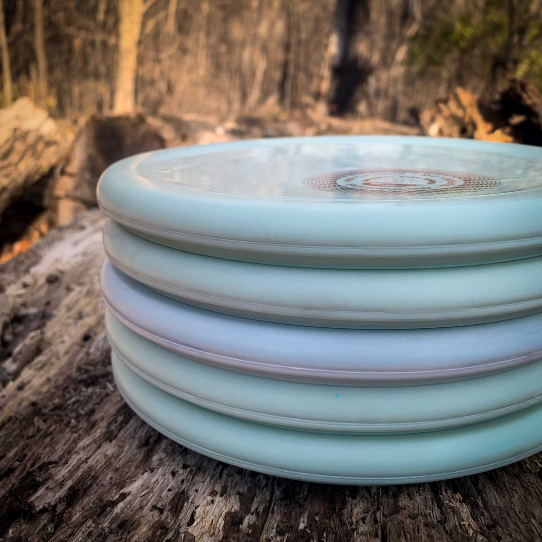 A stack of light blue and white Devilhawk Superglow - SureGrip discs rests on a weathered log in an outdoor wooded area.