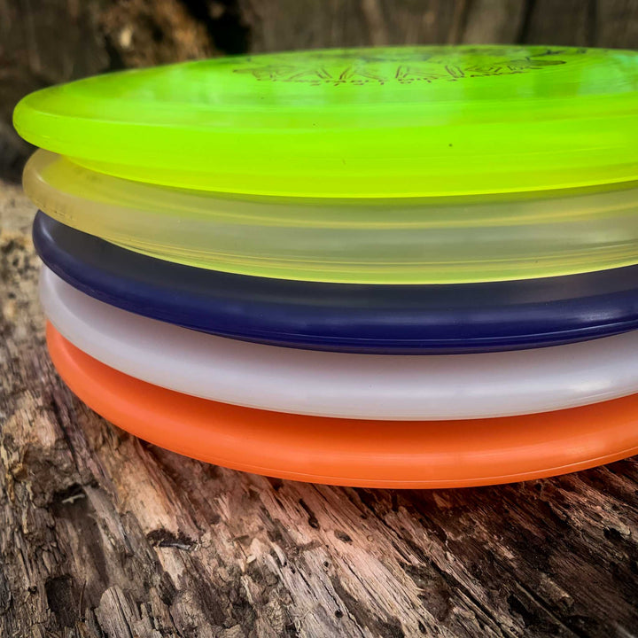 A stack of colorful disc golf discs, including the overstable Warrior - Diamond mid-range disc in green, white, blue, and orange, rests on a rough wooden surface.