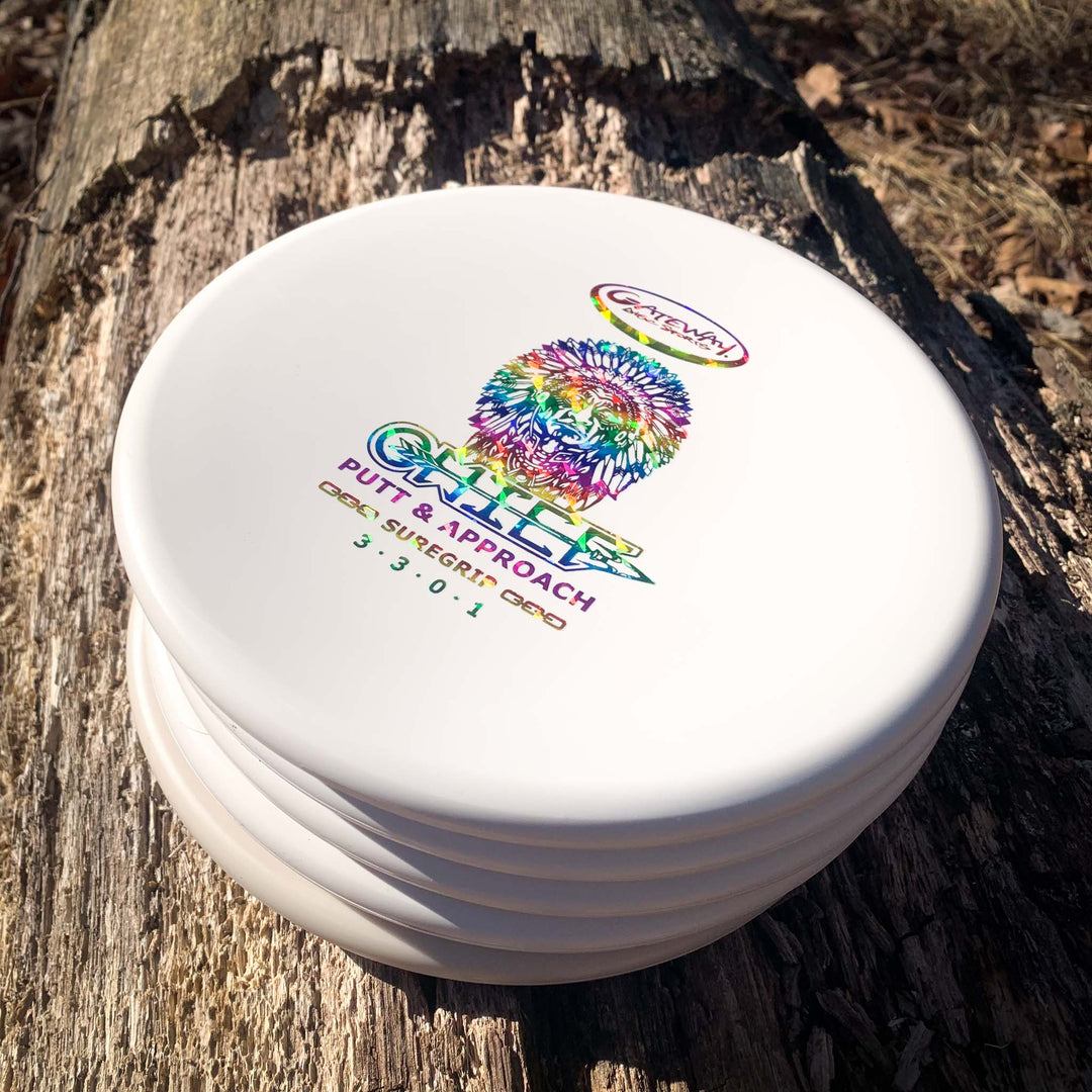 Five white Chief - SureGrip disc golf putters with colorful "Wild" branding and a beaded rim are stacked on a tree trunk in sunlight, ideal for Putt & Approach shots needing extra control.