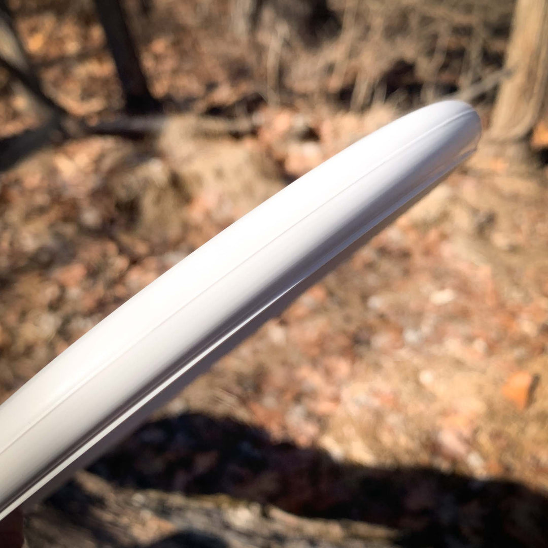 A close-up of the Chief - SureGrip, a white beaded rim flying disc ideal for Putt & Approach shots, held outdoors against a backdrop of blurred trees and dry leaves.