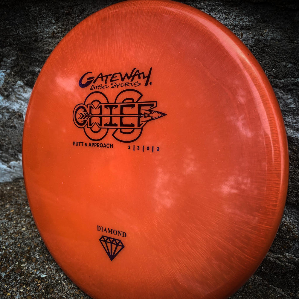 The orange Chief OS - Diamond from Gateway Disc Sports, an overstable putter with black markings and a diamond symbol, rests on rough stone—perfect for controlled hyzer lines.